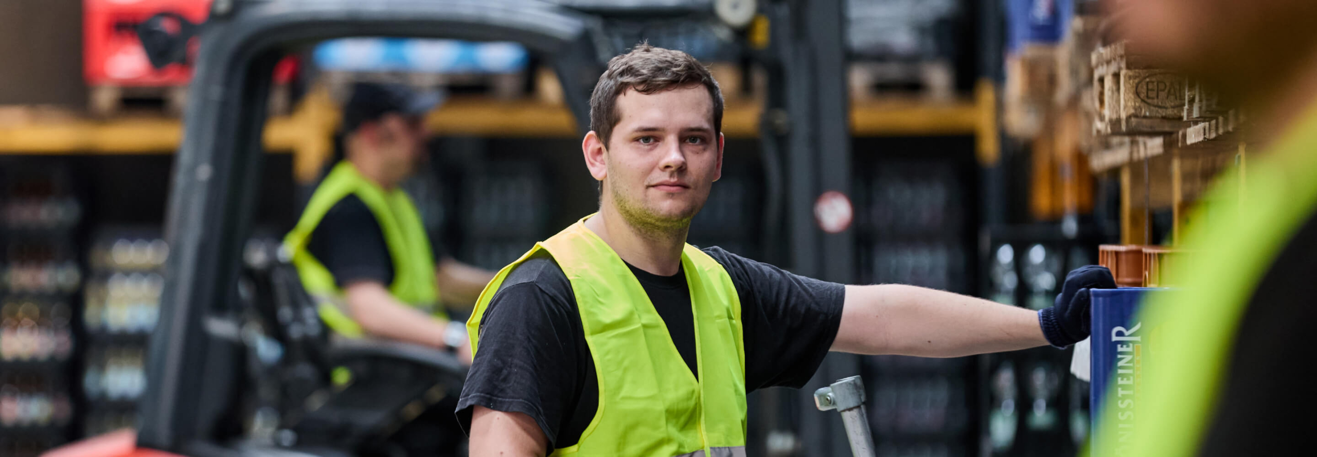 Lagerarbeiter mit Warnweste steht im Getränkelager und hält einen Hubwagen
